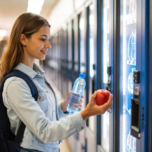 Young woman stands in front of vending machine, holding bottle of water and apple, contemplating her choices. setting is bright and modern, suggesting school or office environment